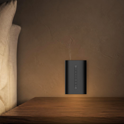 Waterless Diffuser MINI plugged into a bedroom wall above a bedside table, warm amber ambient light and fine dry mist