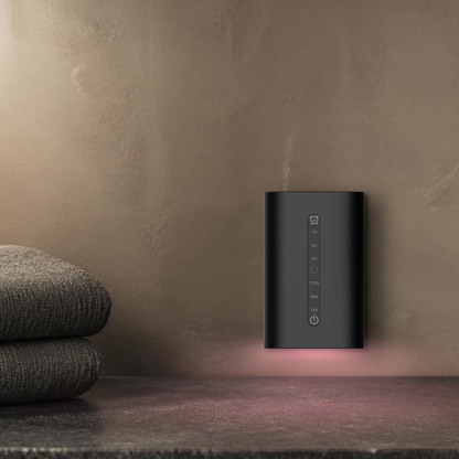 Waterless Diffuser MINI plugged in above a stone bathroom vanity with folded towels, rose ambient light washing the wall